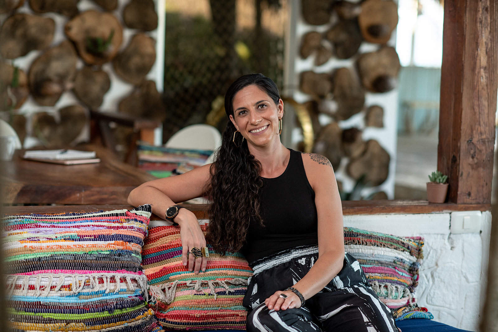 Christine Binko, a white woman and neurodivergent parenting coach, is sitting down facing the camera and smiling. Her arm is resting on vibrant multi-colored striped pillows. She's wearing a black tank top with black and white paperbag bands pants. She has long, dark brown curly hair in a low ponyail and gold hexagon hoop earrings. The background is blurred.