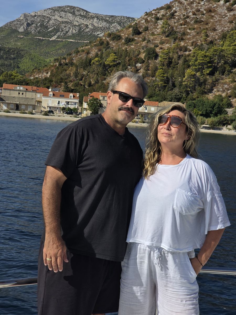 Roy Bryant and his wife, Audra, smiling together on a boat, with blue water, a seaside village, and green hills behind them