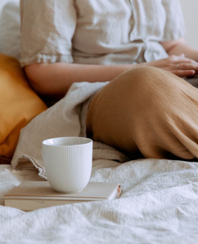 Woman sitting on a bed with a tablet, next to a book and coffee, symbolizing a relaxed approach to simplifying business tech solutions.