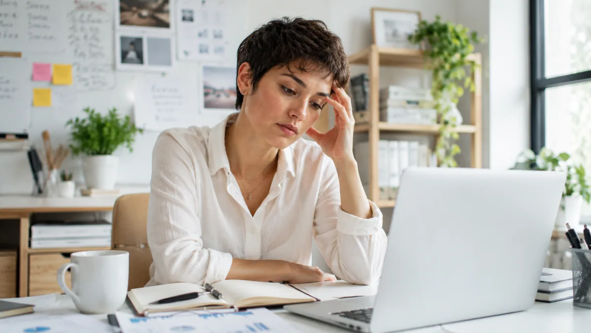 Woman at a bright desk with an open journal and laptop, hand resting on her head, foggy unfocused expression