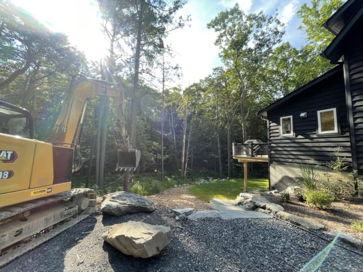 A construction excavator operating next to a dark-colored house in a forest setting, preparing the land for landscaping or further construction.