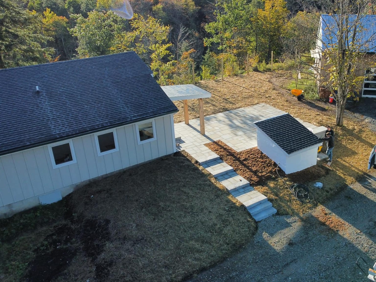 Aerial view of a light-colored house with a small patio under construction, surrounded by mulch, stone walkways, and trees in a rural setting.