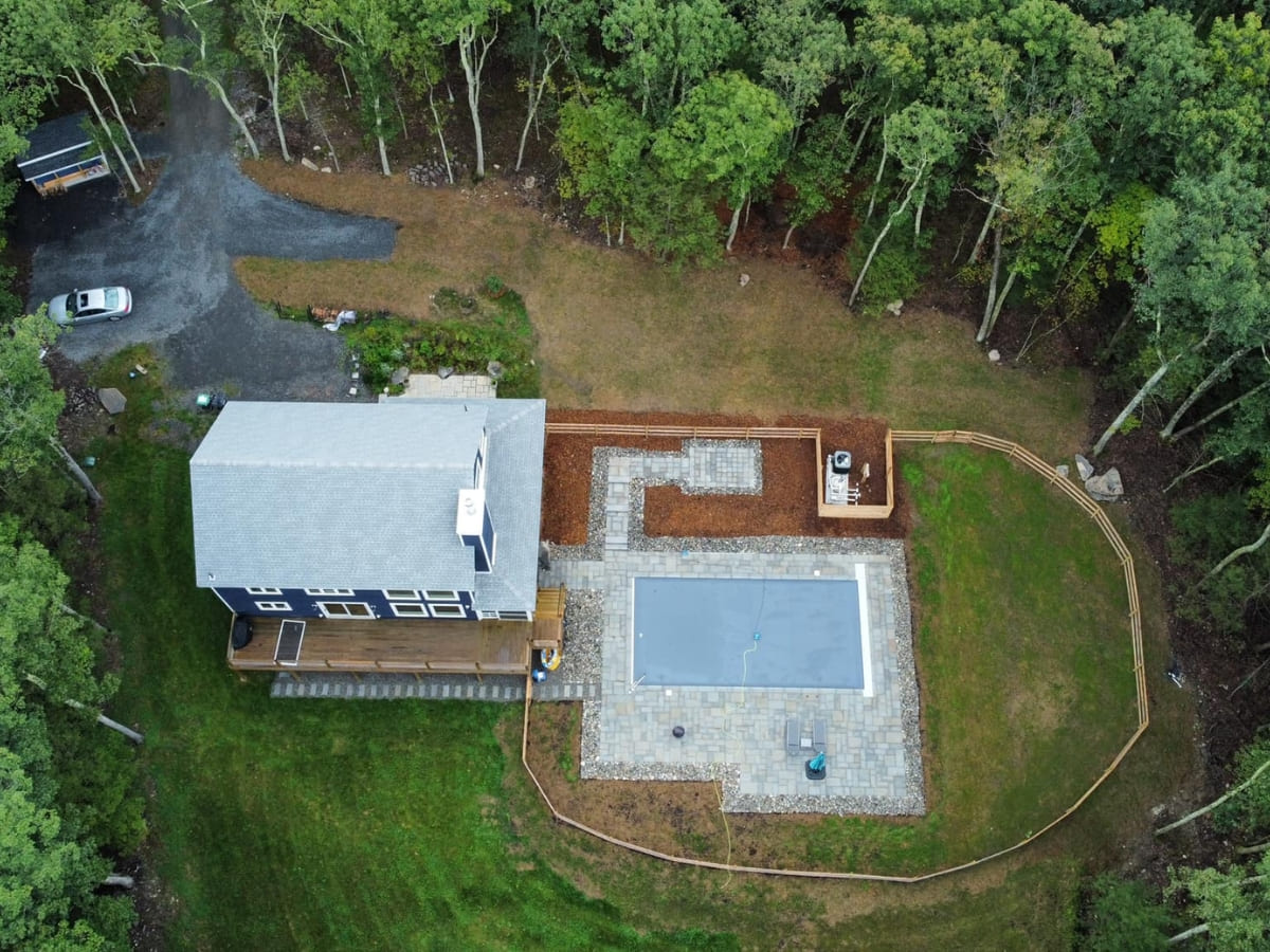 Aerial view of a backyard with a pool, patio, and landscaped garden, surrounded by trees, highlighting a custom home with outdoor living spaces.