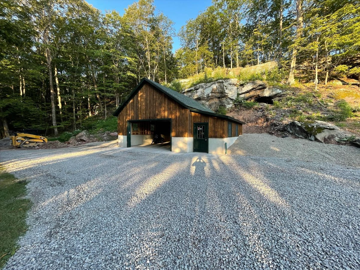 Gravel driveway leading to a large wooden garage surrounded by a wooded area and rocky hillside under bright sunlight.