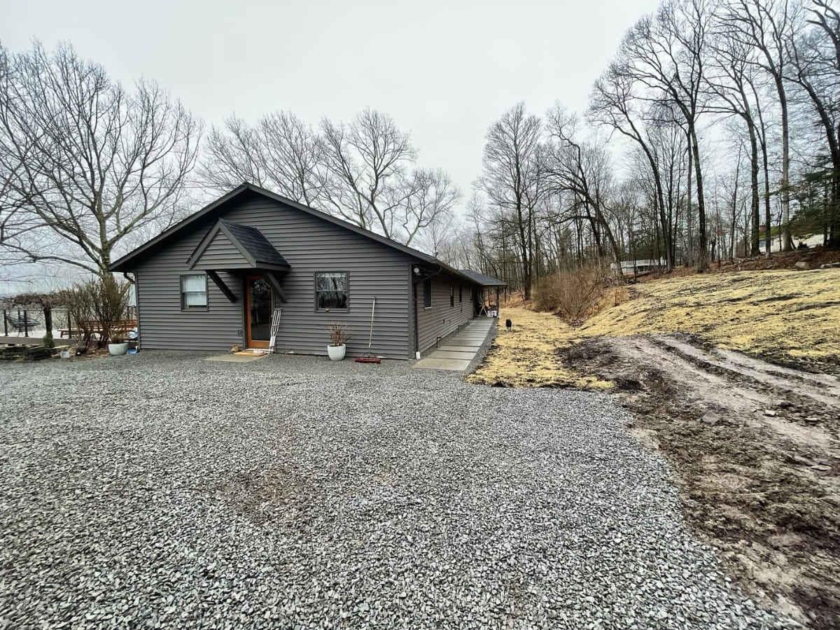 Newly installed gravel driveway in front of a grey house, surrounded by bare trees and sloped landscaping, indicating a completed driveway installation.