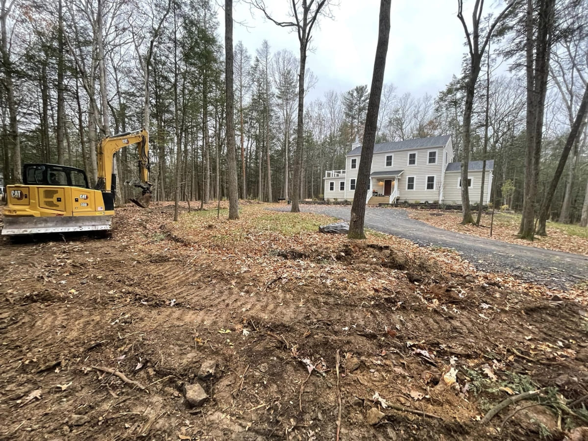 Excavator working in a wooded lot, clearing land next to a large two-story house with a gravel driveway.