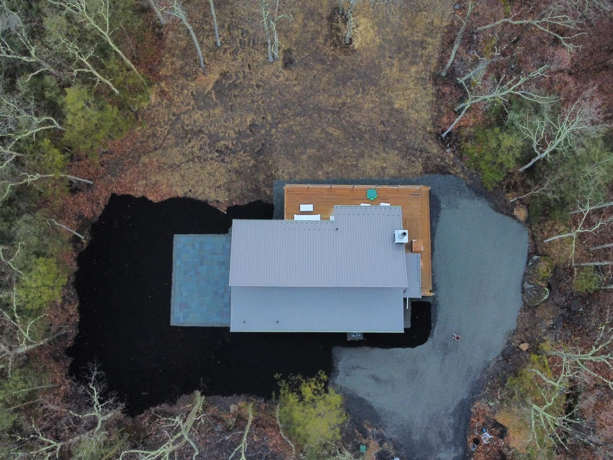 Aerial view of a modern home surrounded by a gravel driveway and dark landscaping, situated in a heavily wooded area.