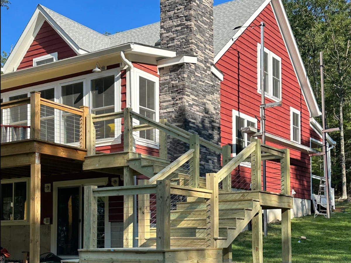 Close-up of a wooden deck and staircase attached to a red house with a stone chimney, featuring modern cable railing.