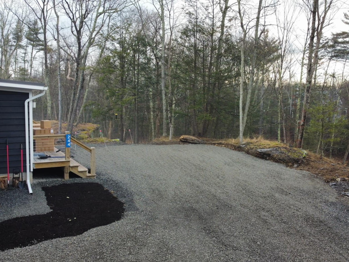 Gravel driveway leading to the side of a dark blue house with a small deck, surrounded by trees and a forested landscape.