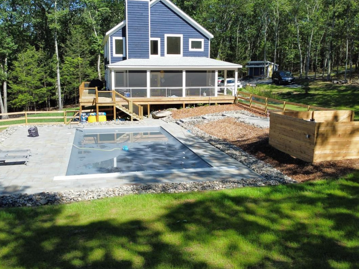 Backyard view of a blue house with a large wooden deck overlooking a newly installed in-ground pool, surrounded by landscaped stone pavers and a garden bed.