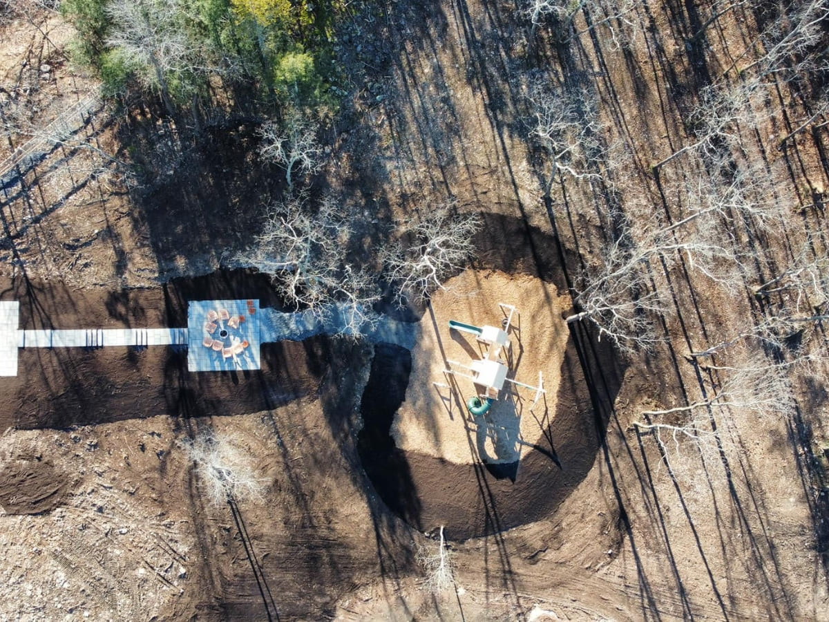 Aerial view of a newly constructed children’s playground and fire pit area in a landscaped backyard, surrounded by dirt and trees.