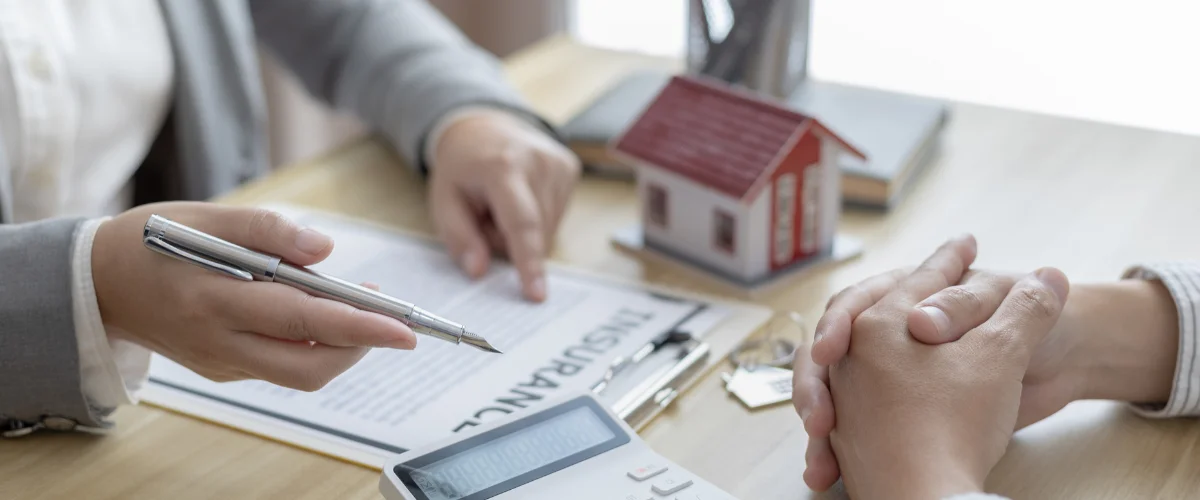 Two people discussing an insurance policy with a calculator, house model, and keys on a wooden table.