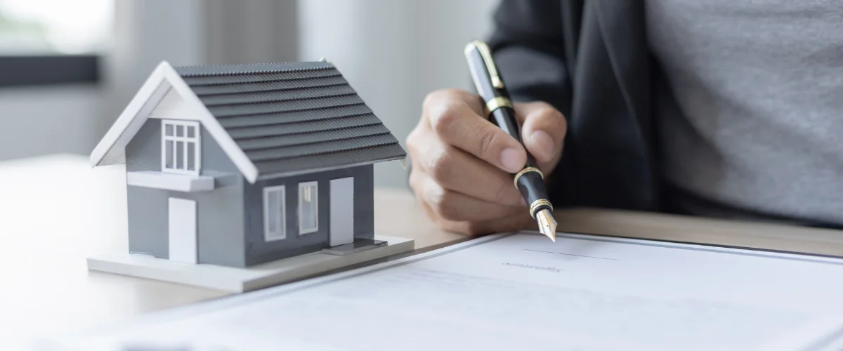 A person's hand with a fountain pen ready to sign a document, with a miniature house model nearby.