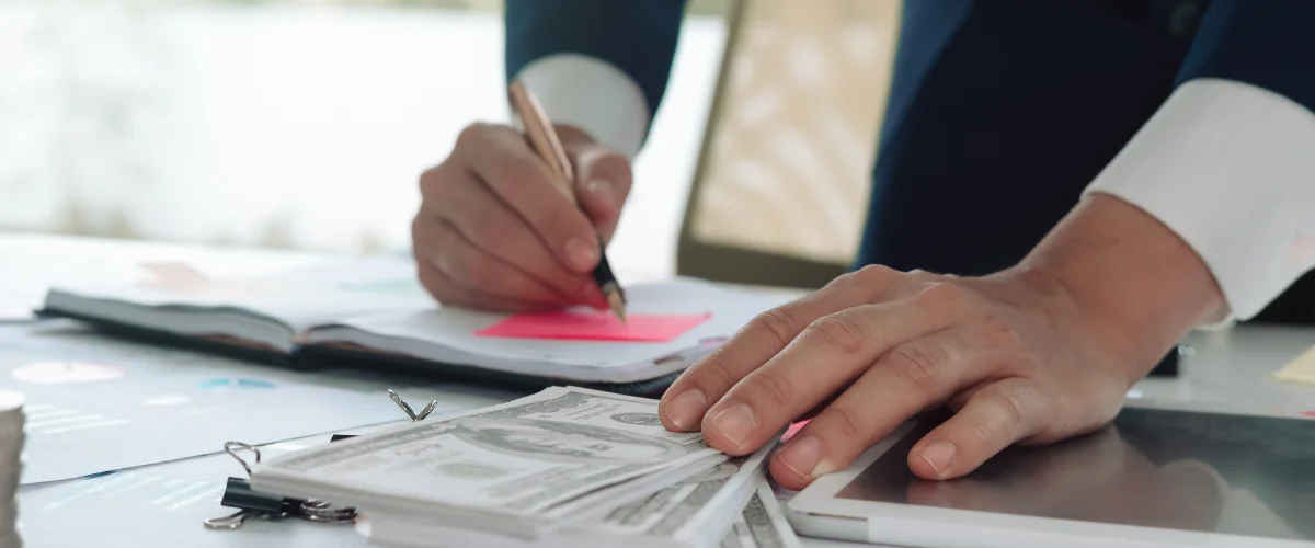 A business person in a suit writes in a notebook, with cash and a tablet on the desk beside them.