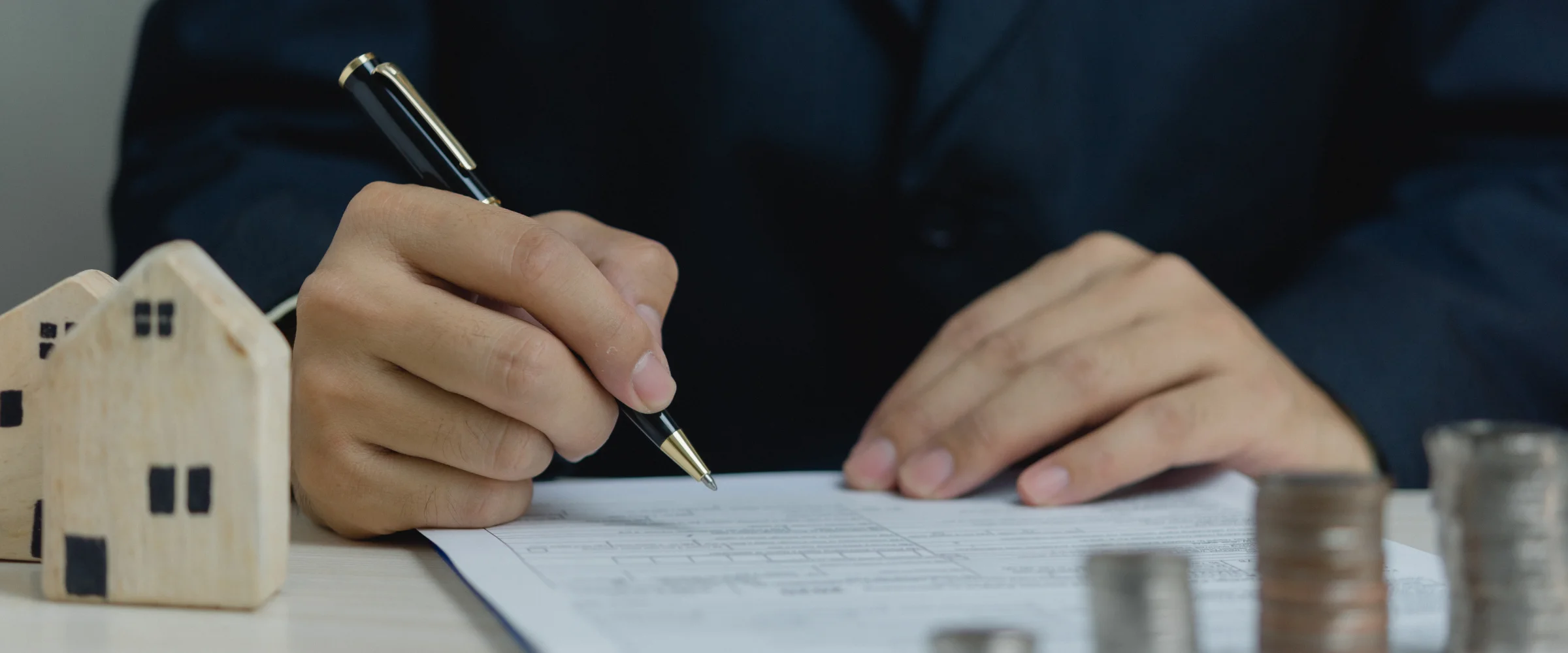 A man's hand in a suit signs a document with a pen, next to miniature wooden houses and stacked coins.