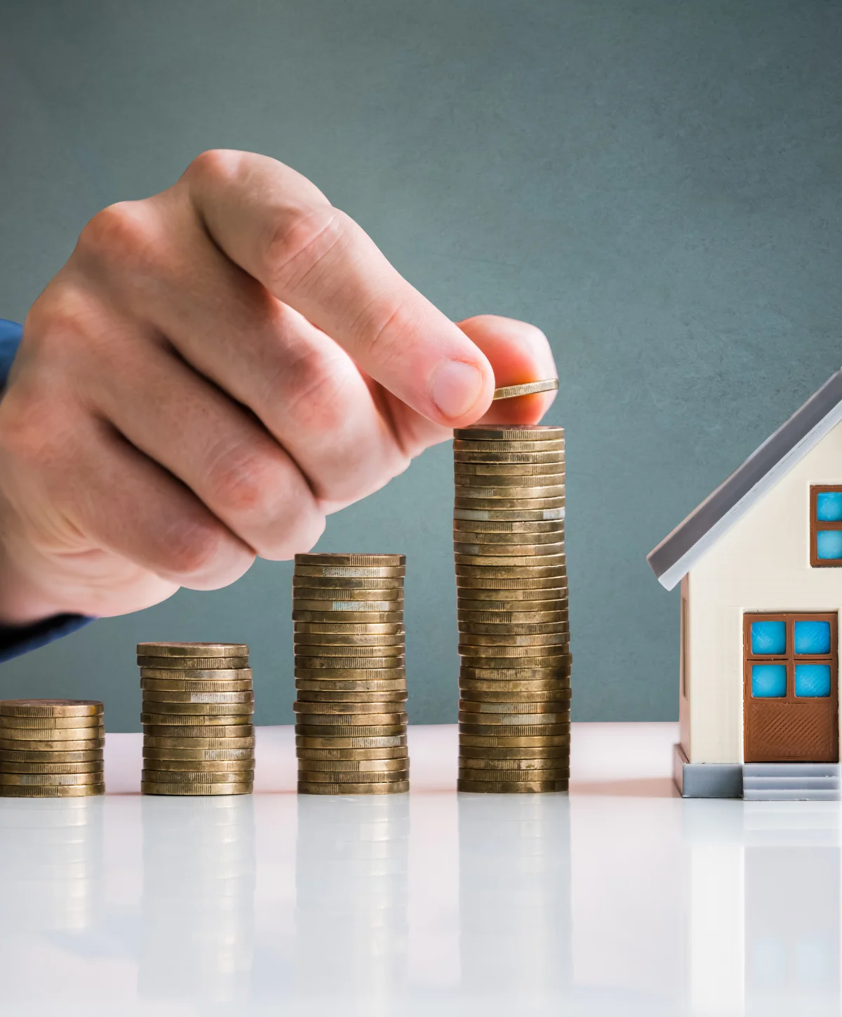 A hand placing a coin on tall stacks of money, with a small house model nearby, symbolizing saving for a home.