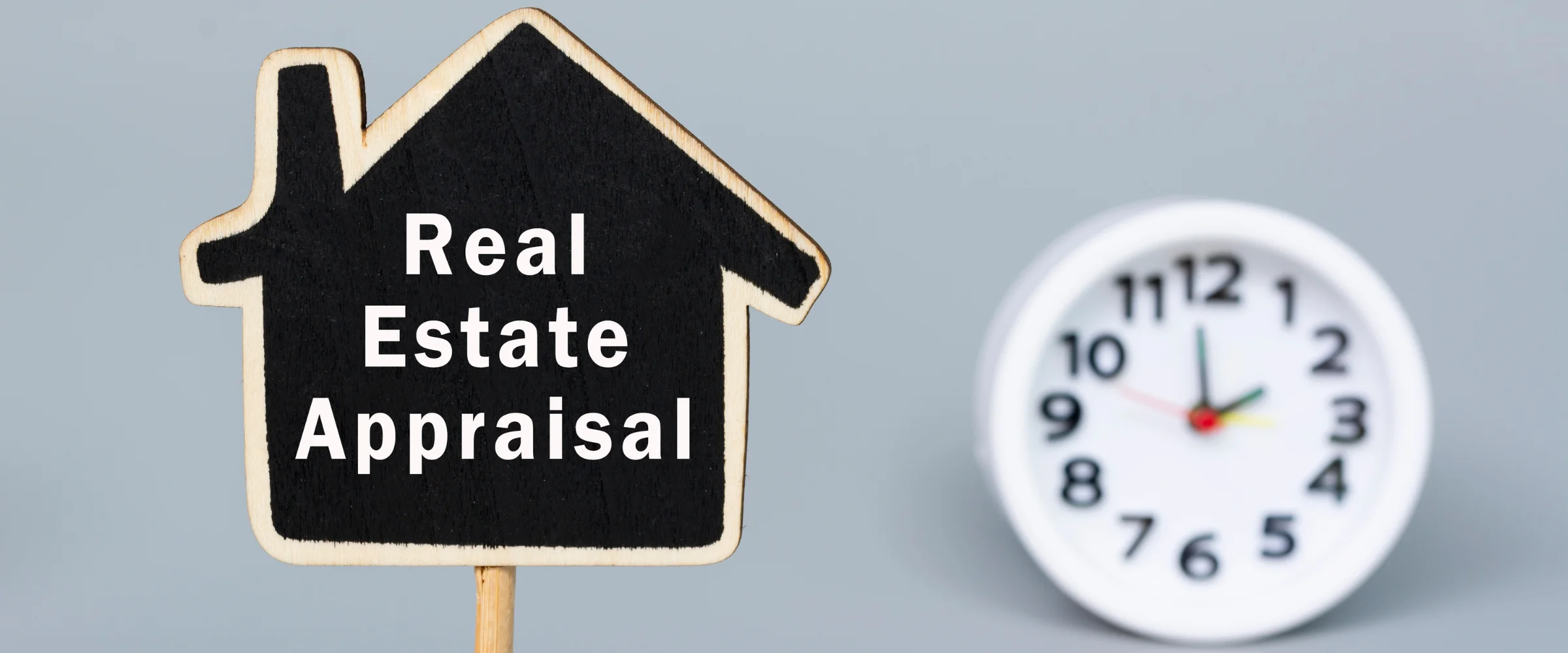 A house-shaped chalkboard sign reading 'Real Estate Appraisal' stands next to a blurred white clock on a grey background.