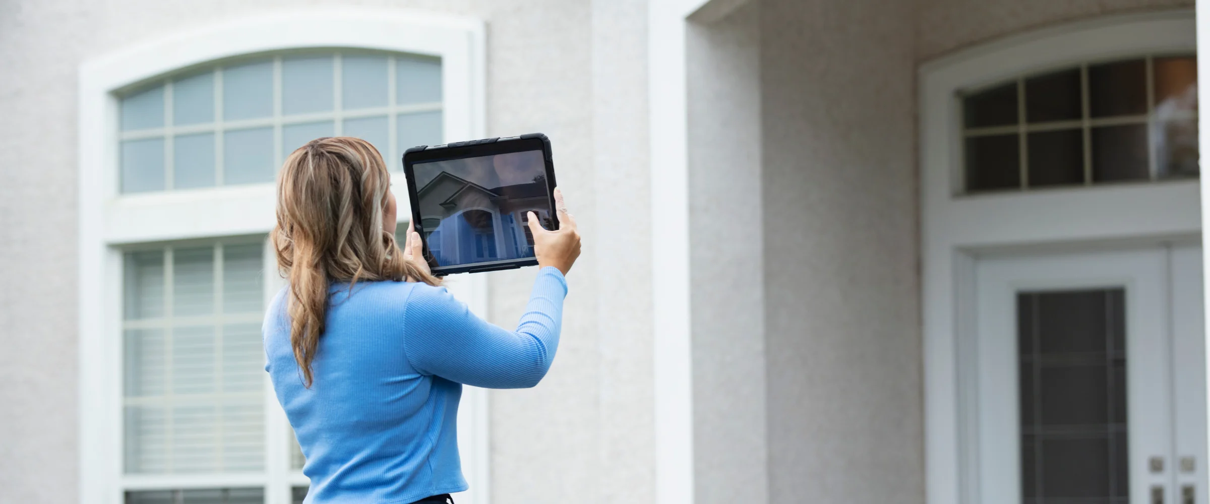 A woman in a blue top records the exterior of a house with a tablet, from a back view.