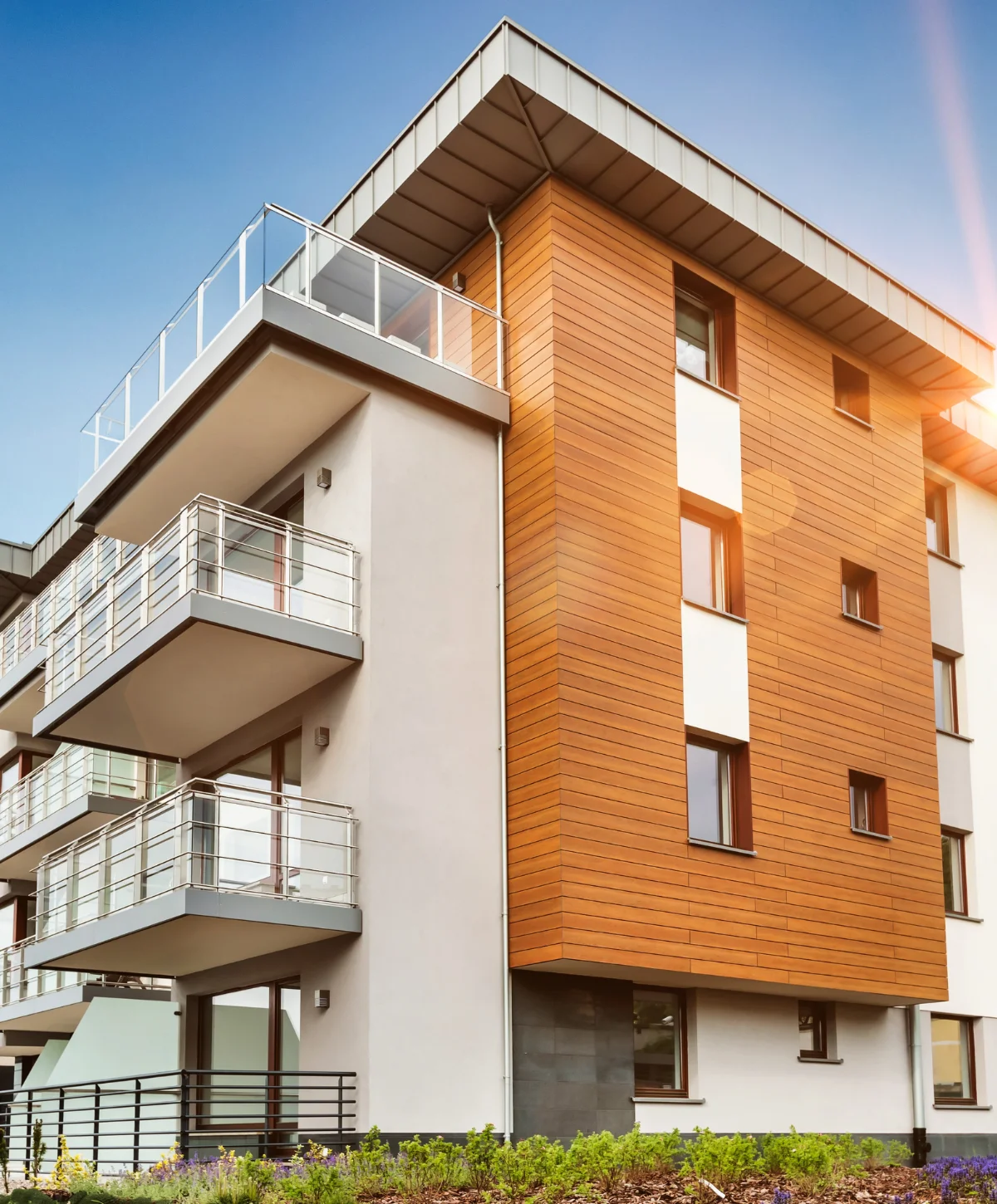 Modern apartment building exterior with warm wooden cladding, light gray walls, and glass balconies under a bright blue sky.