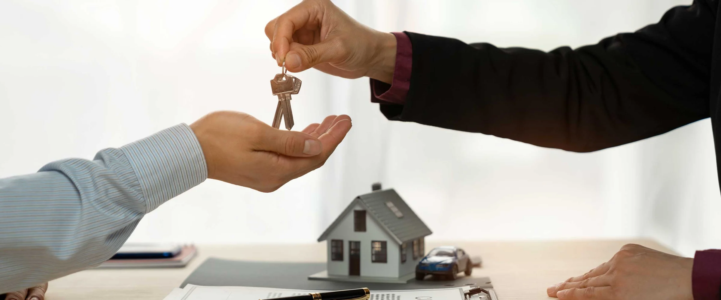 A person in a suit handing house keys to another person, with miniature house and car models on a desk.