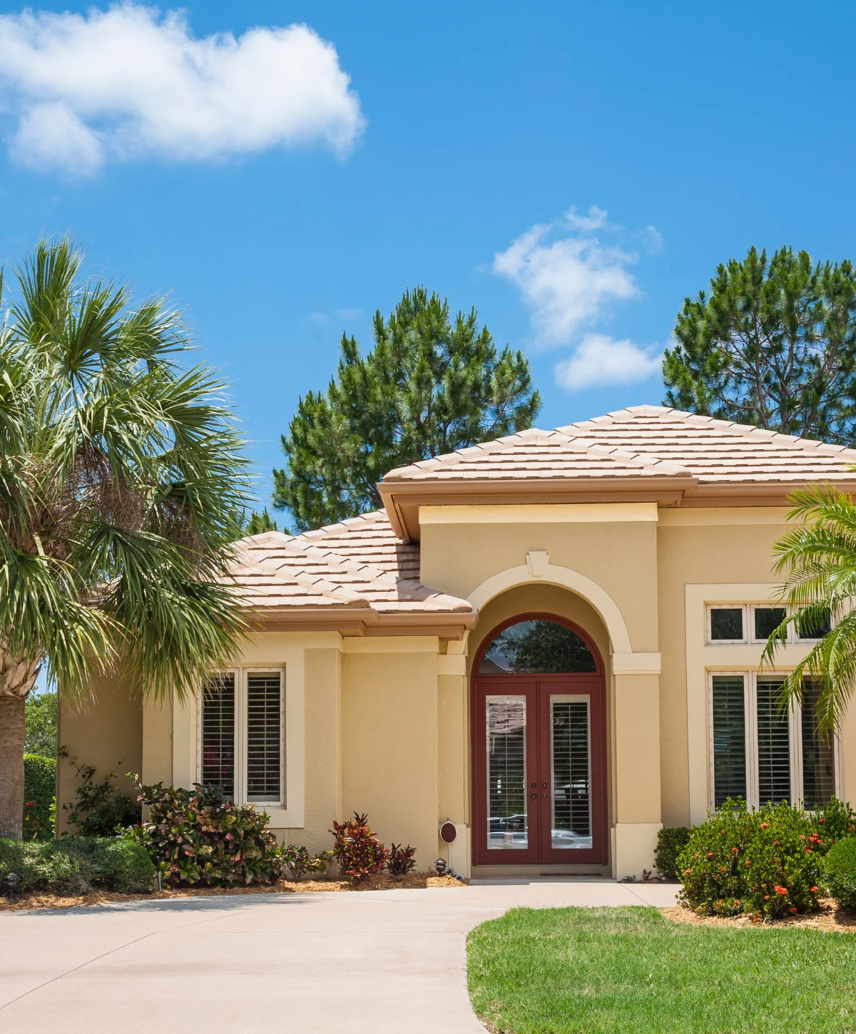 Front view of a light beige stucco house with red double doors, a tile roof, lush landscaping, and a driveway.