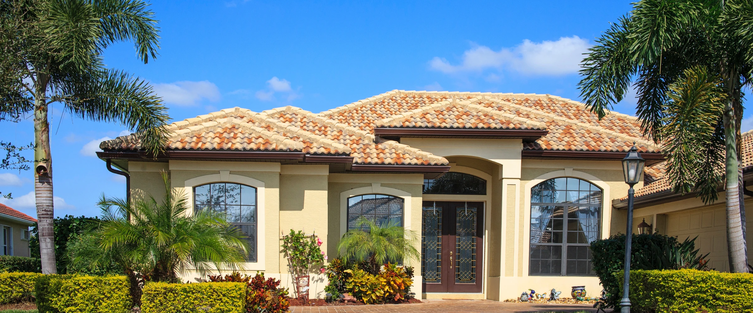 A beige stucco house with a terracotta tile roof, large dark wood entry doors, and arched windows, surrounded by lush tropical landscaping and palm trees under a blue sky.