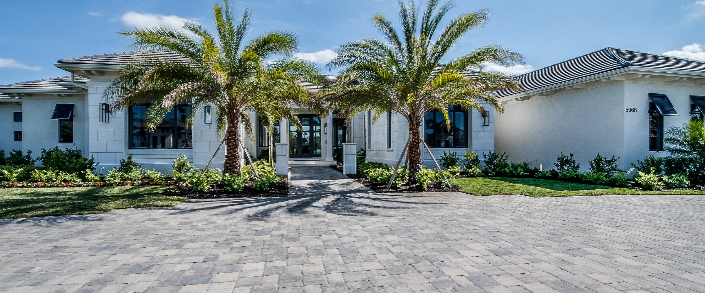 A modern white house with a paved driveway, lush landscaping, and two tall palm trees in front of the main entrance.