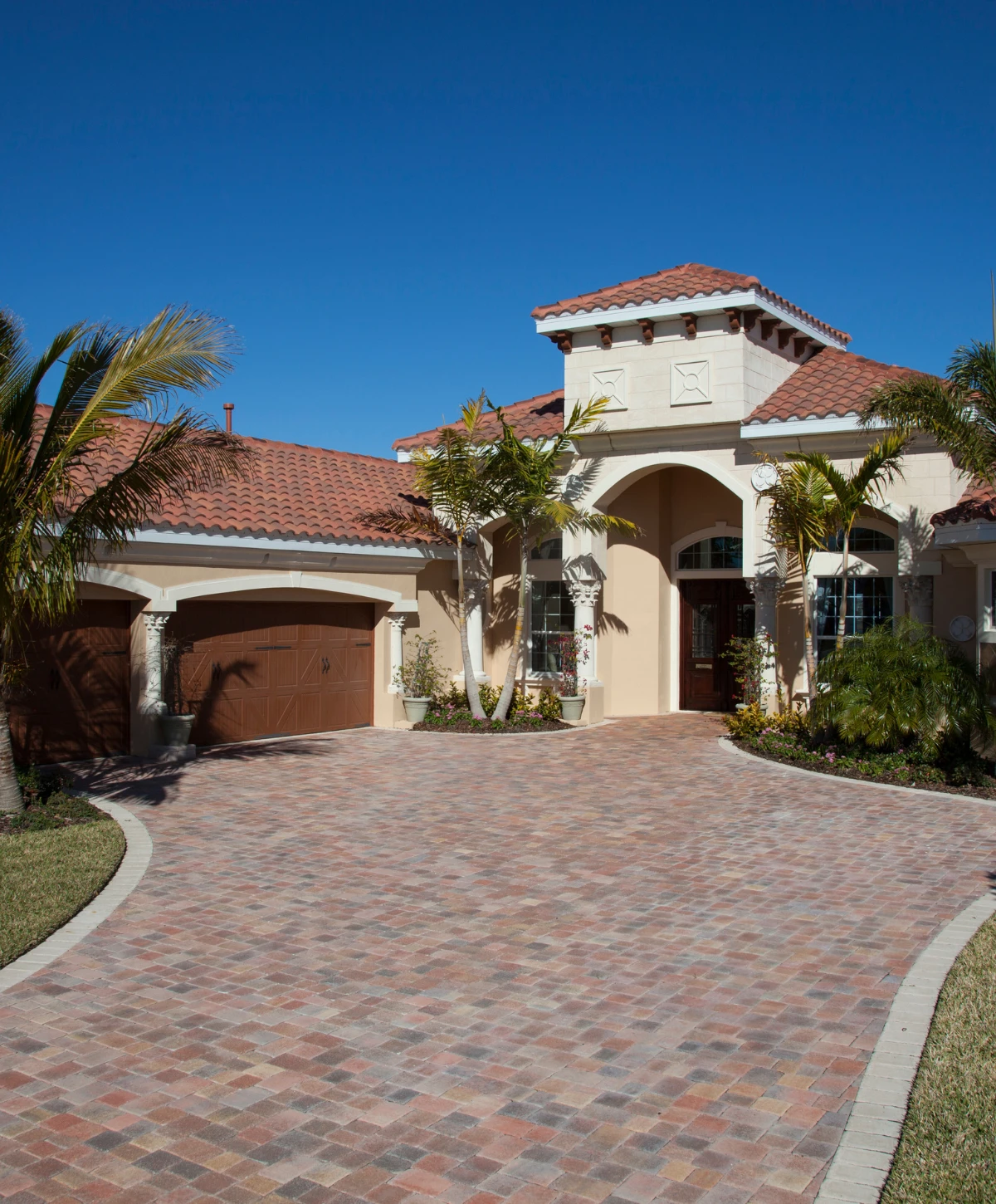A grand Mediterranean-style home with a red tile roof, light stucco walls, and a large multi-colored paver driveway under a clear blue sky.