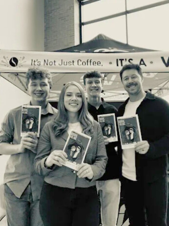 Vibes 2 Go Coffee team holding Midland Community Theatre playbills in front of their V2G vendor booth, showcasing community involvement and local white coffee brand presence.