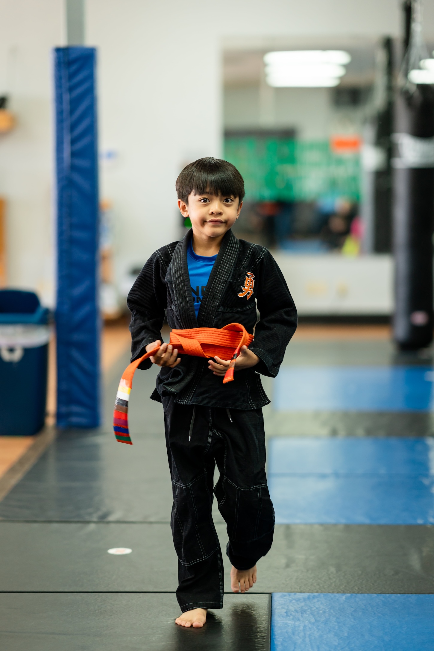 Young boy in a martial arts, walking confidently on mats, wearing a black gi and holding an orange belt..