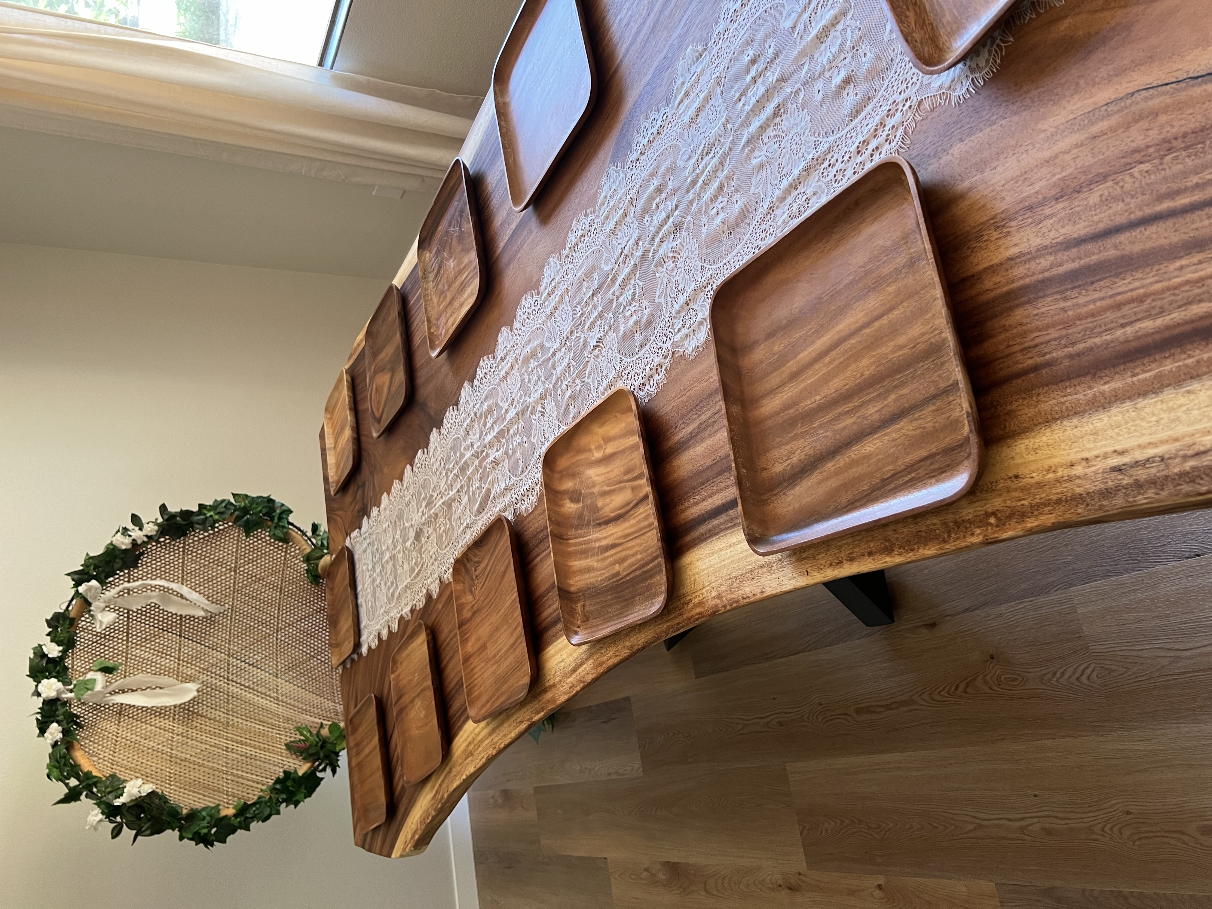A long live-edge wooden table set with a white lace runner and square wooden plates, ready for guests.