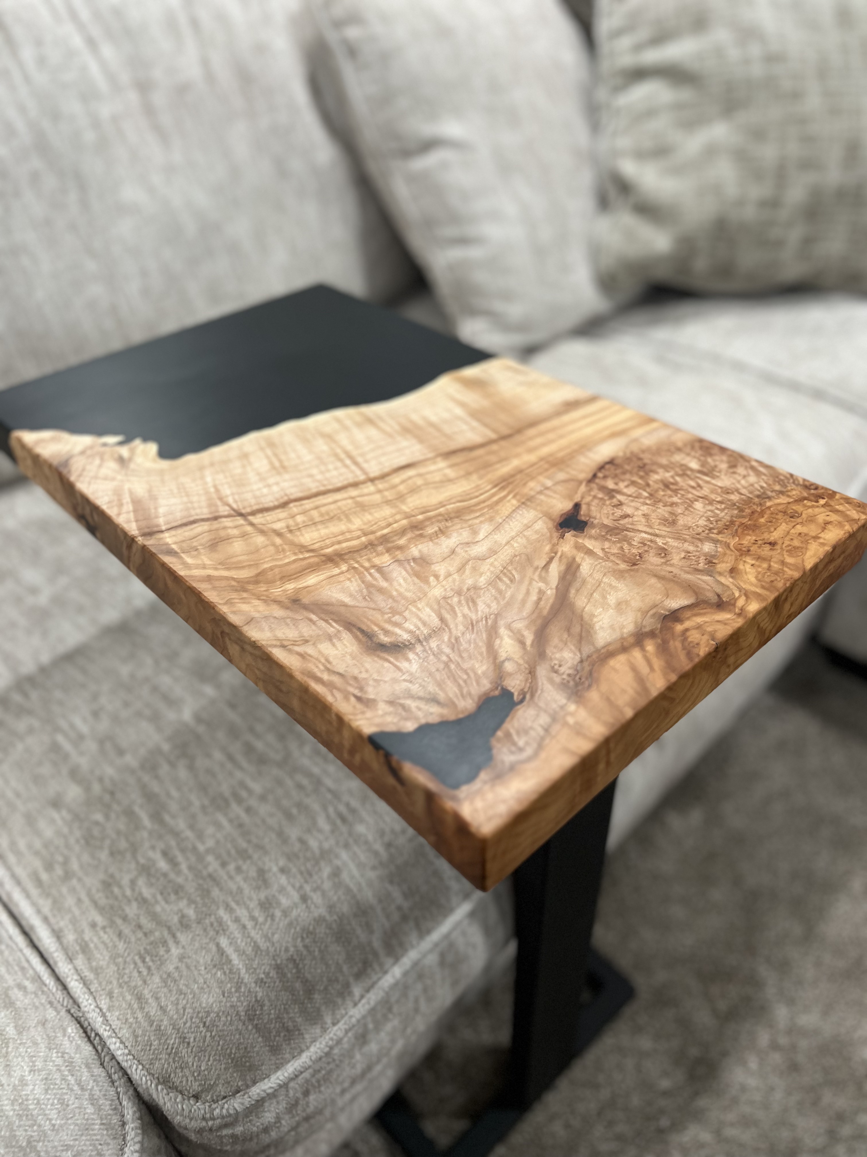 A modern side table with a light live-edge wood top and black epoxy resin inlay beside a grey sofa.