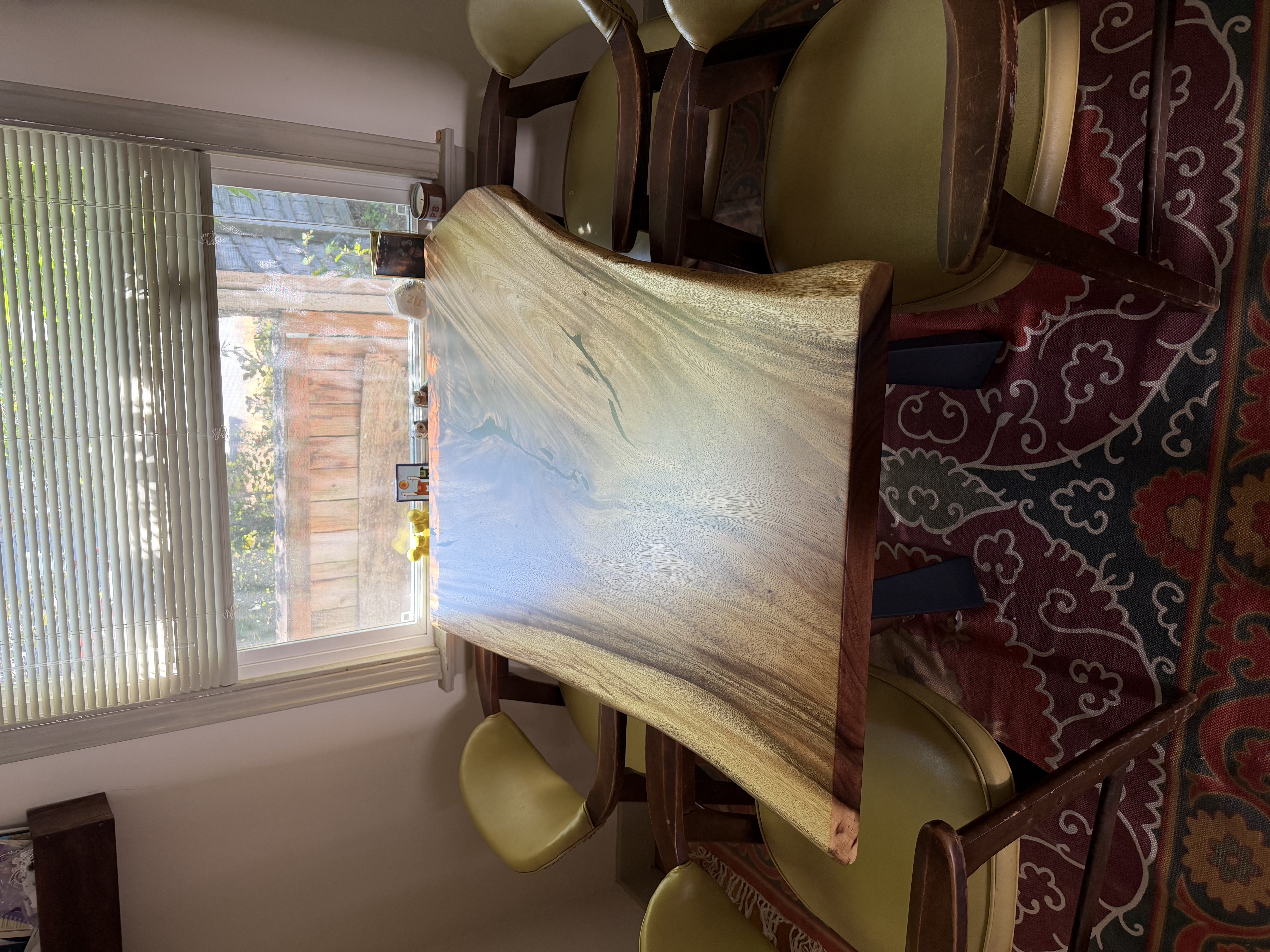 A live-edge dining table with a subtle blue epoxy inlay, surrounded by green mid-century chairs by a window.