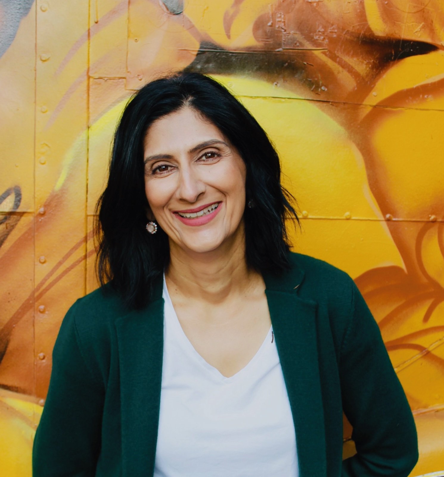 Salena Kulkarni in a green jacket, smiling, in front of a yellow mural