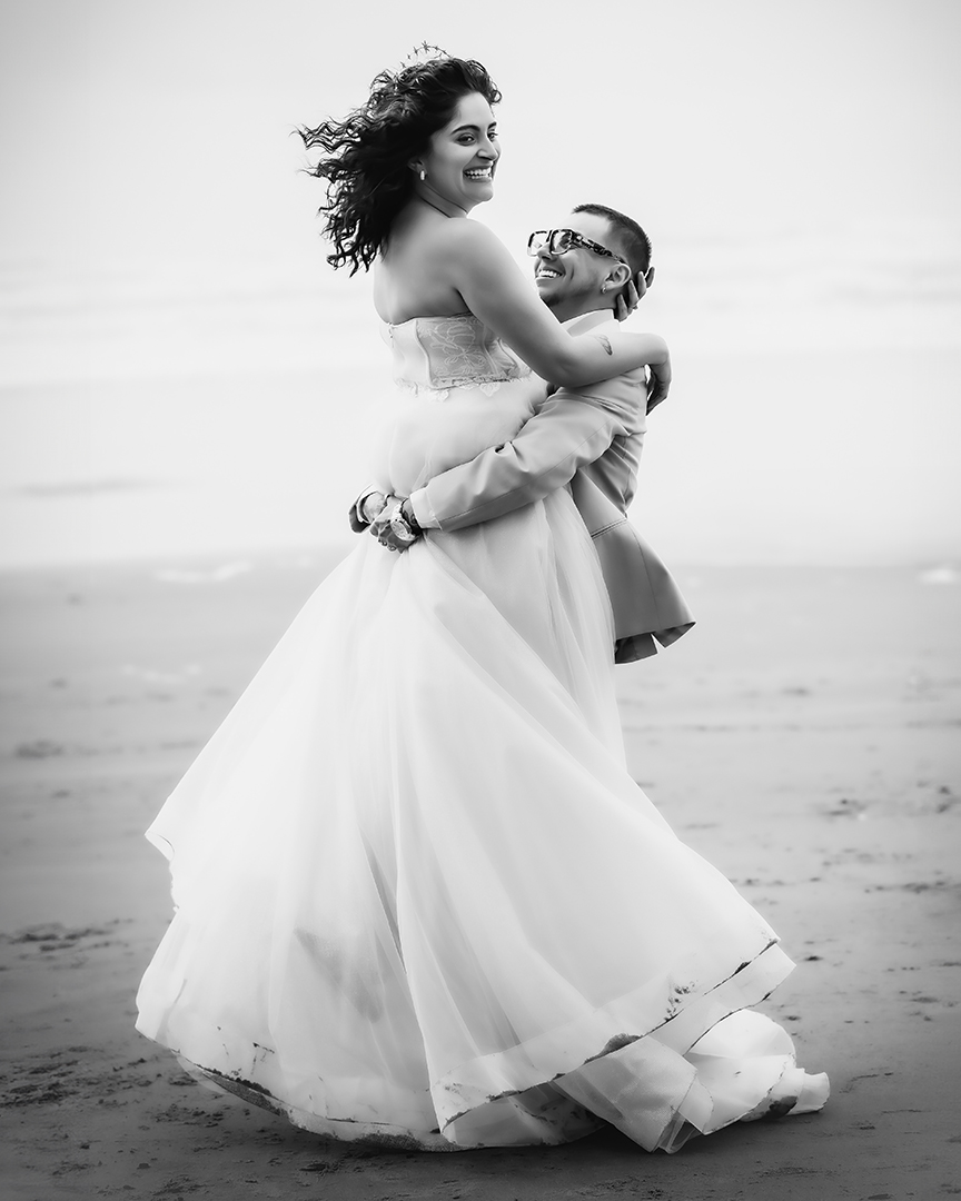 A couple is on a beach near Seattle The groom lifts his bride and twirls her around in a circle. Both have smiles and the groom is wearing glasses. 