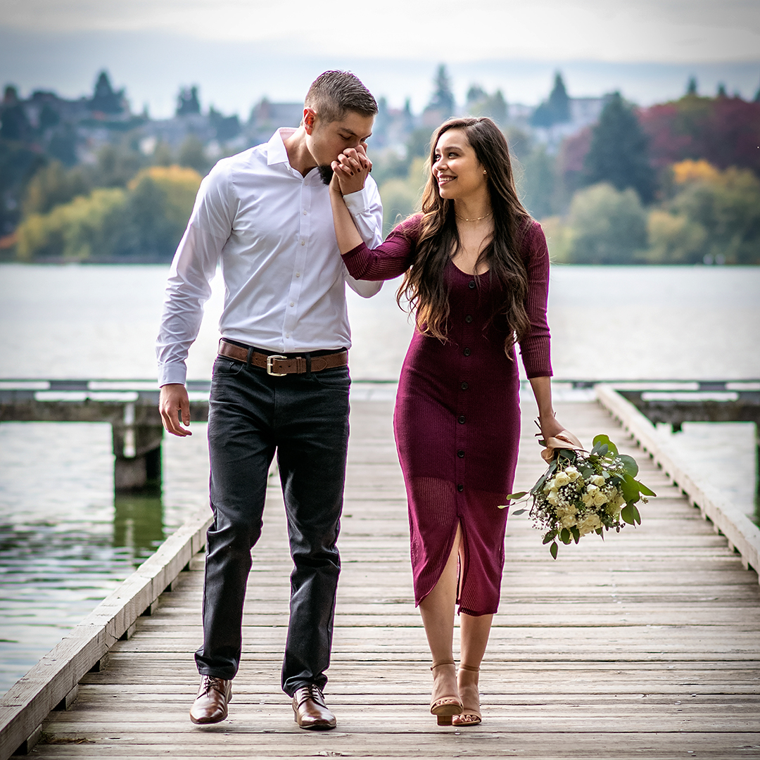 Couple kissing whie standing on a mossy rock in front of a waterfall. The man is wearing a tan jacket with black jeans and brown boots. The woman is wearing blue jeans and a white sweater with white tennis shoes. Her blond hair is blowing in the wind from the spray of the waterfall. 