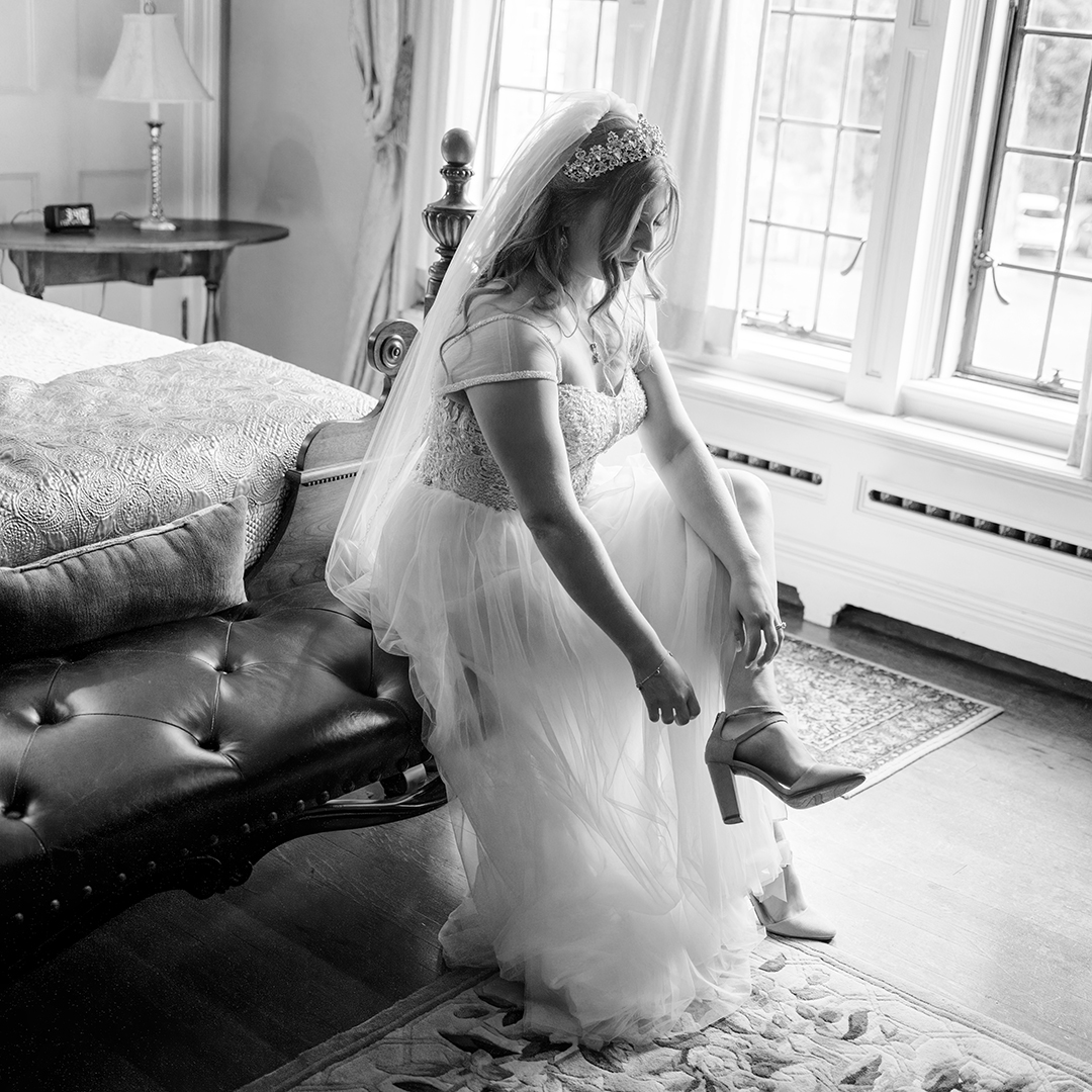 Bride putting on her shoes while getting ready for her wedding day. She's wearing heels and sitting on a bench at the foot of a bed. 