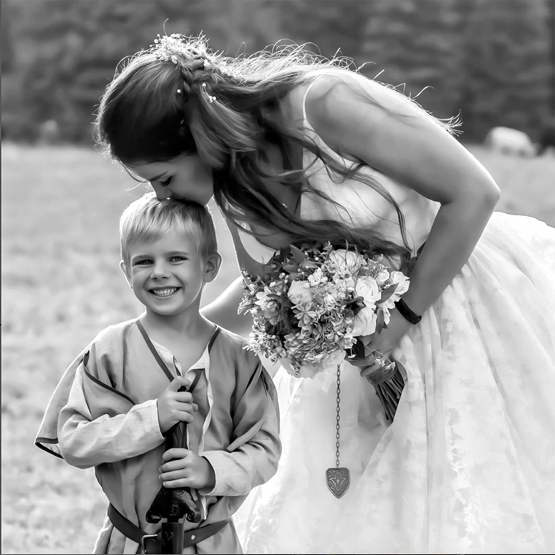 Bride holds bouquet of flowers while kissing little boy on the top of the hea. Boy is holding a sword and carried the wedding rings as the ring bearer. They are standing in a forest meadow. 