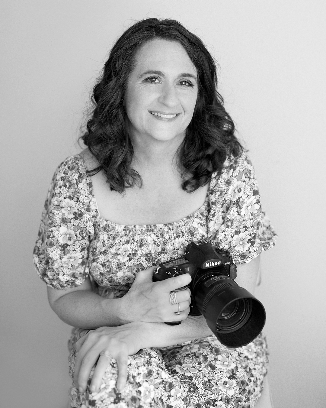 A black and white photo of the photographer holding her camera sitting on a stool. 