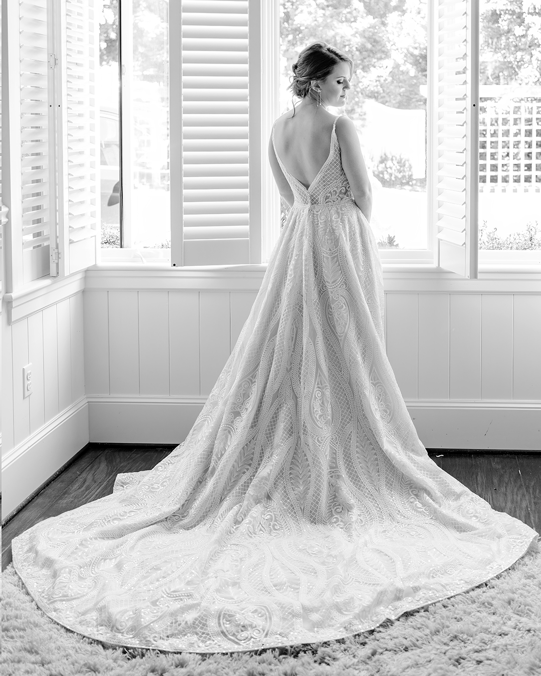 Bride standing at a window with shutters over window slighty open. Bride is looking over her shoulder in a white lace gown with her eyes closed. 