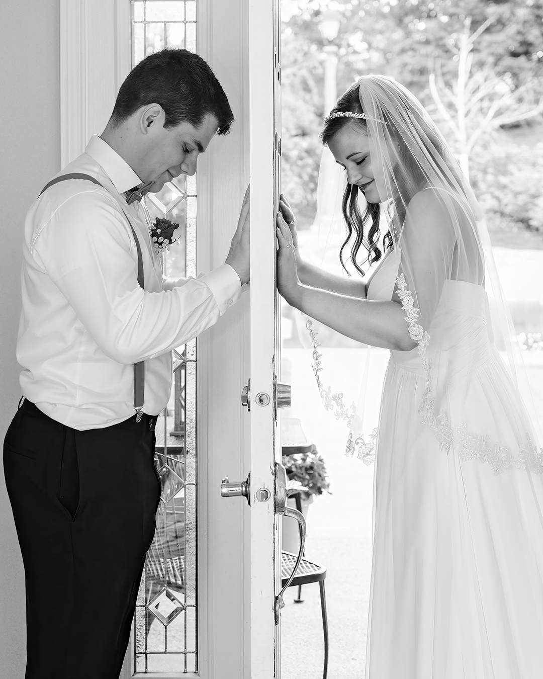 A Couple Stands on either side of the door for their first look in Seattle. The photo  in black and white. The bride has a waist length veil and the groom is wearing a white shirt with suspend