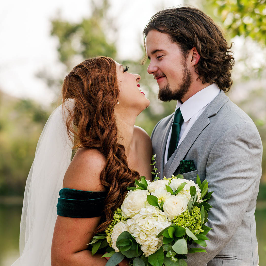 A couple stands close together with big smiles on their faces. The bride is wearing an emerald green edding dress with a cathedral length veil and holding a floral bouquet of white chrysanthemums and green leaves and florals. The groom is wearing a grey suit with an emerald green tie and pocket square.