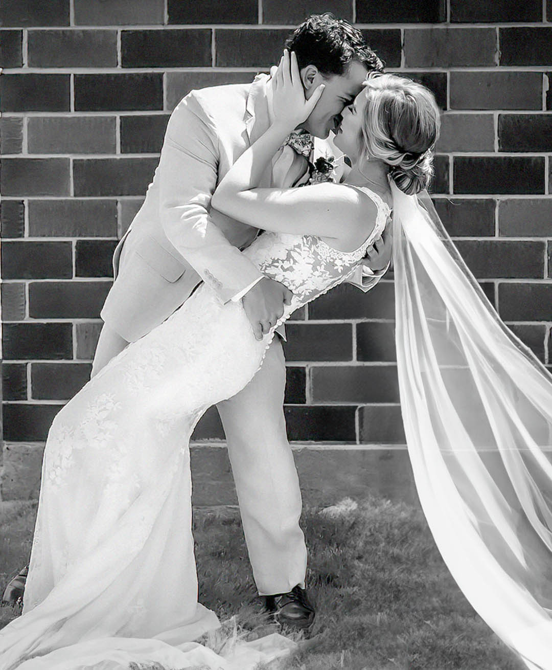 A couple cuddles during their first look giving a smiling kiss and dip during their private first look. The photo is in black and white and the bride as a low bun with a long veil. 