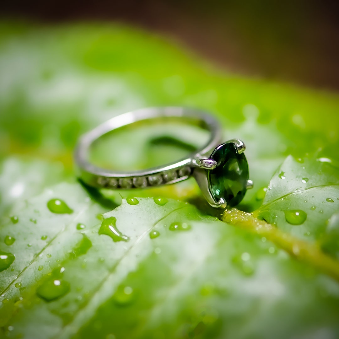 Emerald green wedding ring laying on a green leaf with rain drops sprinkled on the leaf. 