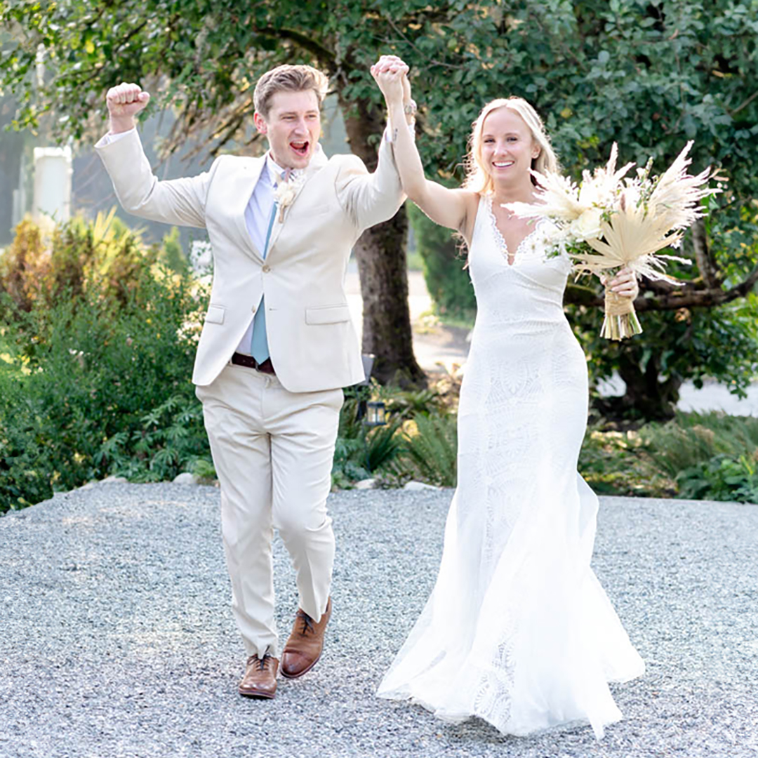 Bride and groom cheer as they enter the reception during their wedding day. Bride is holding a bouquet of flowers and groom is wearing a tan suit. 