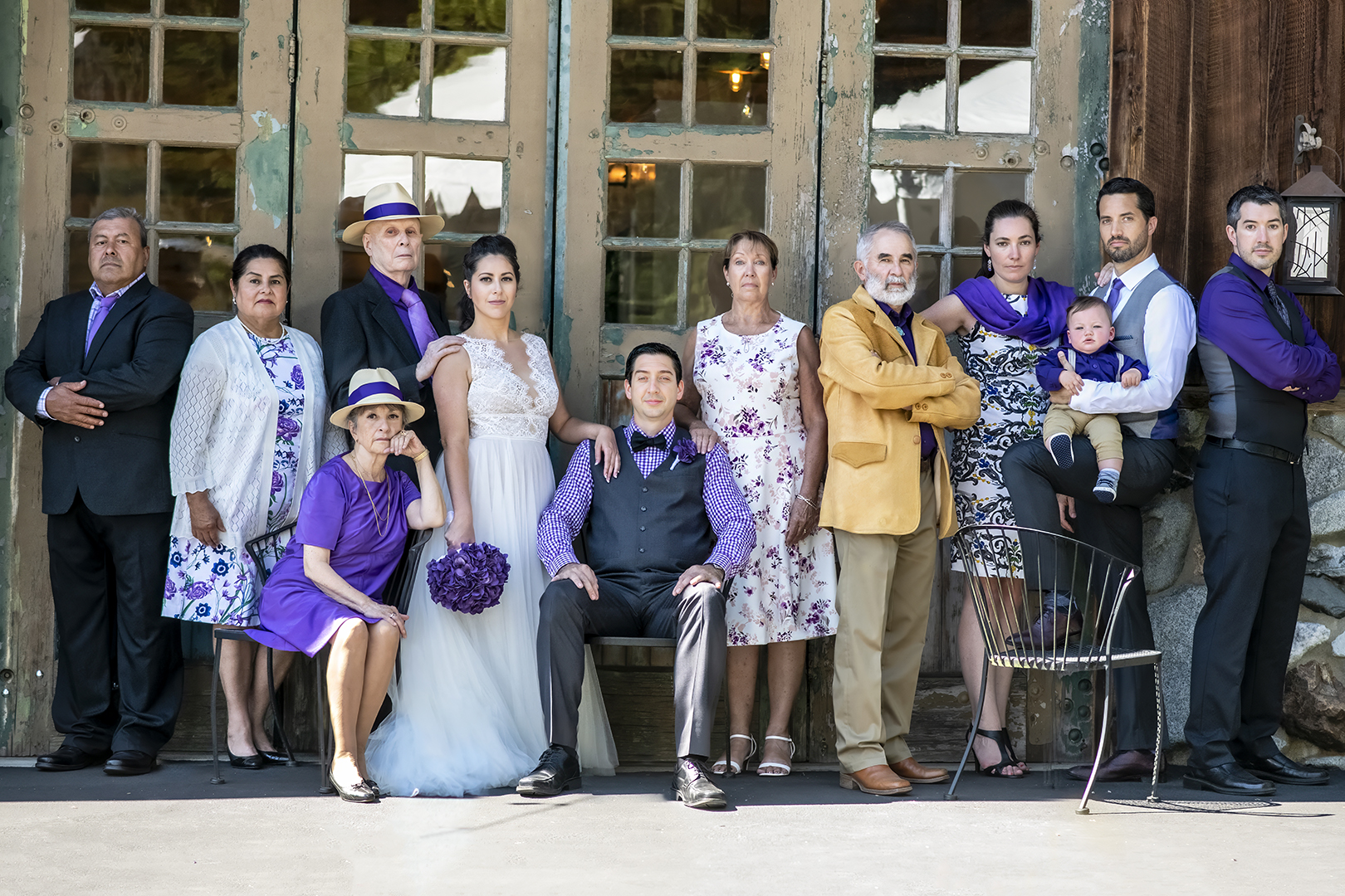 Dynasty setting family shot on a wedding day. The family is standing in front of old fire station doors that has paint chipping off them. The groom is sitting along with the mother of the bride. The bride is standing next to the groom with her hand on his shoulder. 