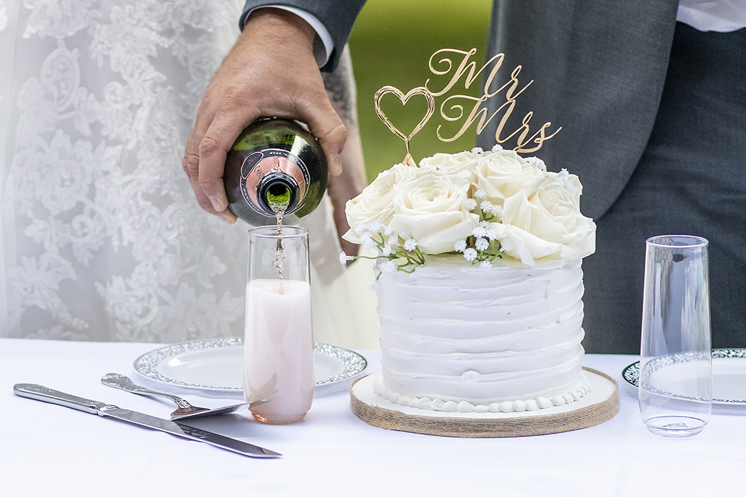 Groom pours a glass of pink champagn next to their white wedding cake. 