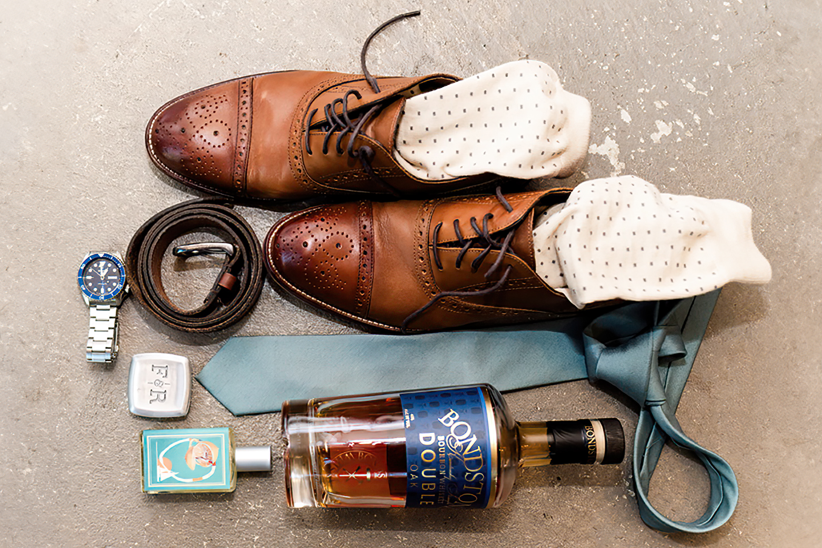 Grooms detail items on a wedding day which include: his shoes, socks, belt, watch, necktie, bottle of whiskey, and cologne.