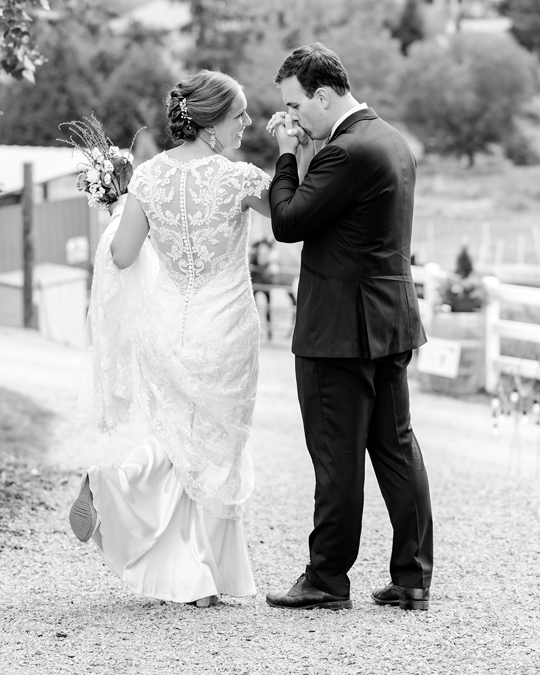 Bride and groom walking on a path at their wedding venue. Groom kisses the bride's hand while she looks affectionately at the groom. The bride is holding the train of her dress and her bouquet of flowers. Groom is wearing a black tux. 