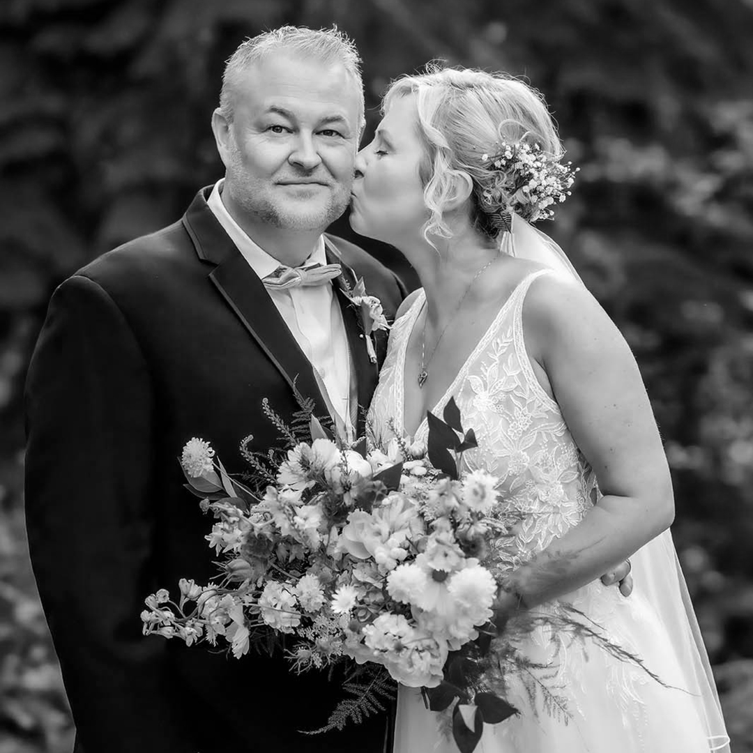 Bride kisses the Groom's cheek while holding a bouquet of blue and white flowers. Bride is wearing a lace wedding gown with her hair in a low bun and a veil tucked into the bun. The groom is wearing a black tuxedo with a blue bow tie. 